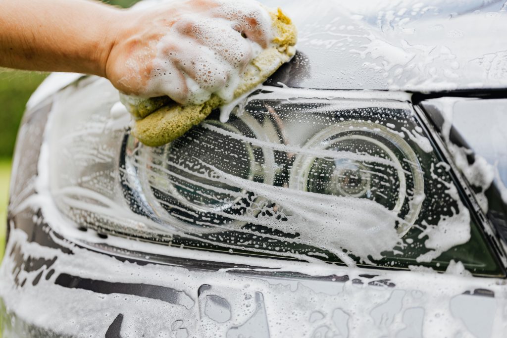 Hand scrubbing a car headlight with sponge and soap suds, detailing process.