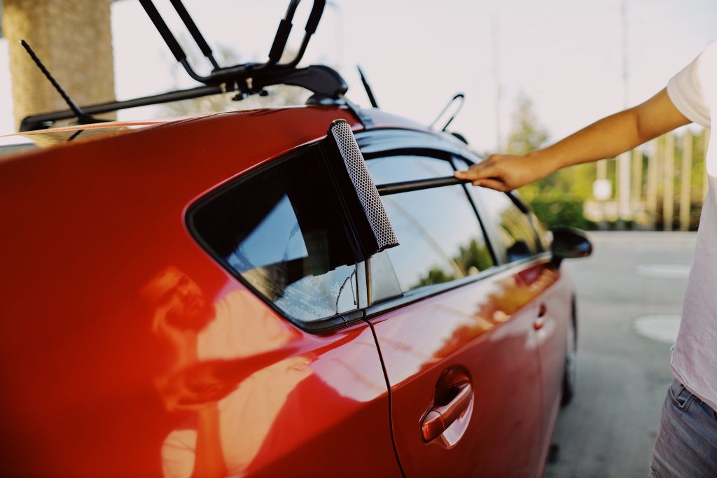 Close-up of a person using a squeegee to clean a red car window outdoors.